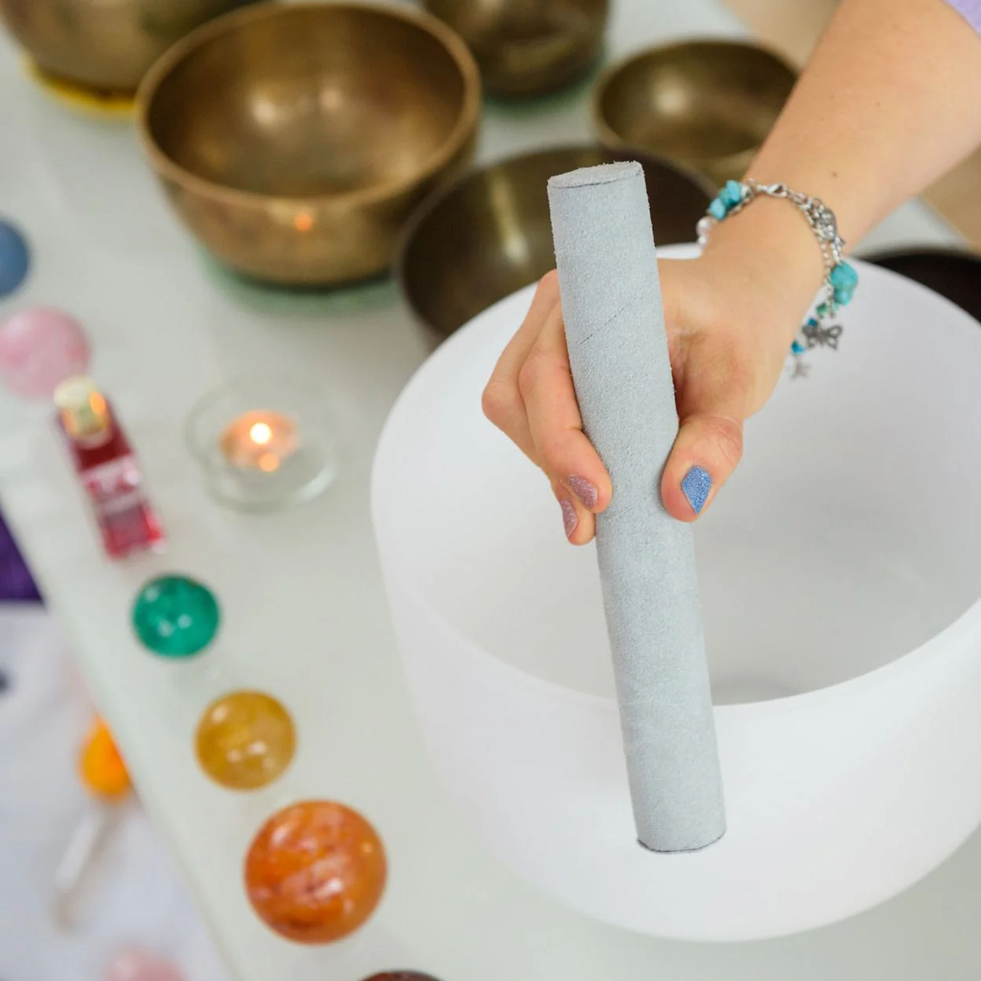 Hand holding a suede mallet over a white crystal singing bowl, surrounded by brass bowls, colorful crystal spheres and a lit tealight — sound healing meditation setup in a wellness studio.