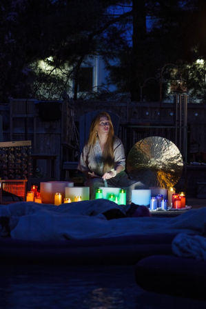 Nighttime backyard sound bath with a person seated among glowing multicolored candles, crystal singing bowls and a large gong, cozy blankets in the foreground