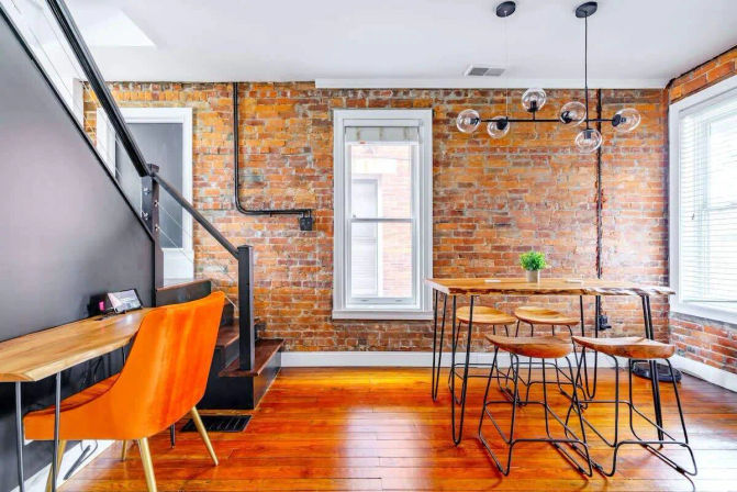 Urban loft dining nook with exposed brick wall and hardwood floors; live-edge high-top wooden table with metal stools, modern globe pendant light, bright orange accent chair by a staircase and large windows.