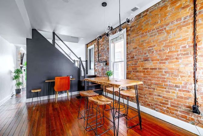 Cozy industrial-style loft dining area with exposed brick wall, live-edge wood table, metal-legged stools, hardwood floors, and hanging globe pendant lights.