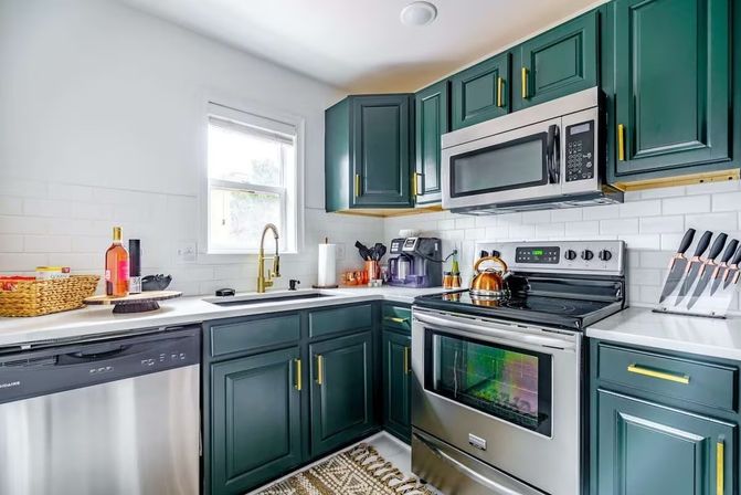 Sunlit modern kitchen with deep green shaker cabinets, white subway tile backsplash, gold hardware and faucet, stainless steel stove, microwave and dishwasher, white countertops and patterned rug