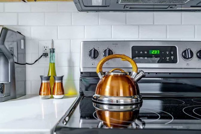 Gleaming copper tea kettle with wooden handle on a black glass electric stovetop in a modern kitchen, stainless steel range, white subway tile backsplash and countertop oil bottle and spice shakers