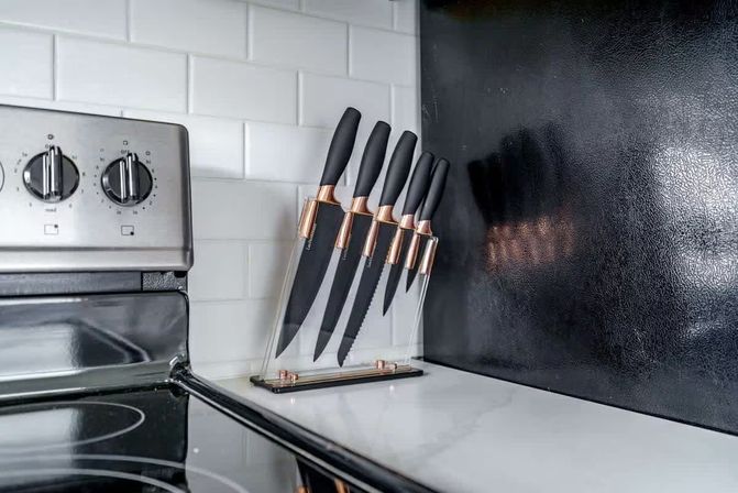 Sleek modern kitchen countertop with a clear acrylic knife block holding black-handled knives with rose-gold accents beside a stainless-steel stove and white subway-tile backsplash.