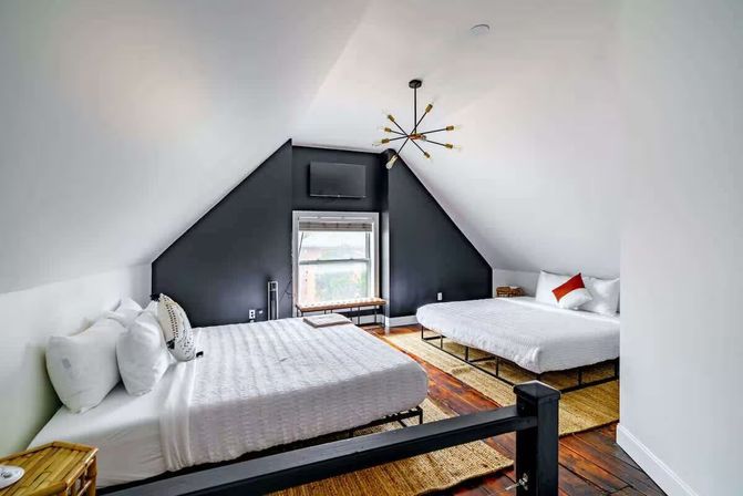 Cozy attic loft bedroom with two queen beds in white linens, dark triangular accent wall, sputnik chandelier, hardwood floors, woven rugs and a window bench.