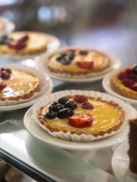 Close-up of glossy fruit-topped custard tarts with blueberries and sliced strawberries on white plates in a bakery display case.