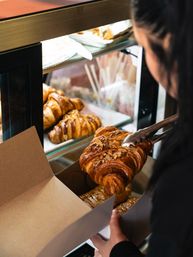 Almond-topped croissant picked up with tongs and placed into a takeaway box at a bakery display case with golden croissants behind it
