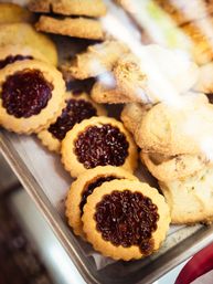 Close-up of a bakery display tray with golden jam-filled thumbprint cookies and crumbly shortbread biscuits, glossy red berry jam centers.