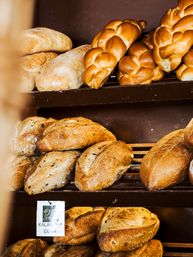 Warm golden braided challah and rustic artisan loaves, including Kalamata olive sourdough, stacked on wooden bakery shelves