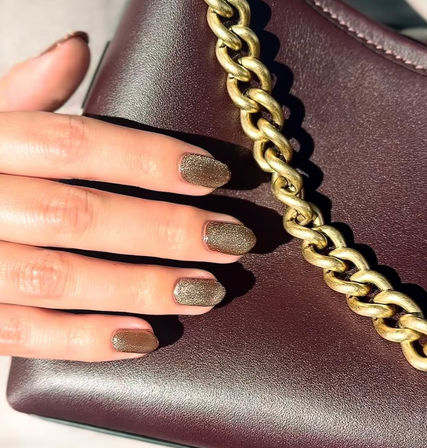Close-up of a hand with textured gold glitter manicure resting on a burgundy leather handbag with a chunky gold chain — stylish nails and purse detail.