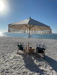Sunny beach scene with a fringed umbrella shading two striped lounge chairs and a small side table on white sand, calm blue ocean and swimmers on the horizon.
