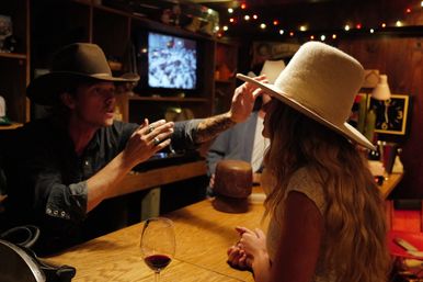 Man in a dark cowboy hat adjusts a woman’s oversized cream felt hat across a cozy wooden bar counter, with a glass of red wine and string lights overhead.