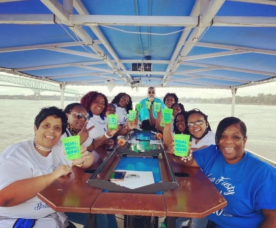 Smiling group on a covered pedal‑bar river cruise raising neon green cups around a wooden bar table with a bridge and waterfront in the background