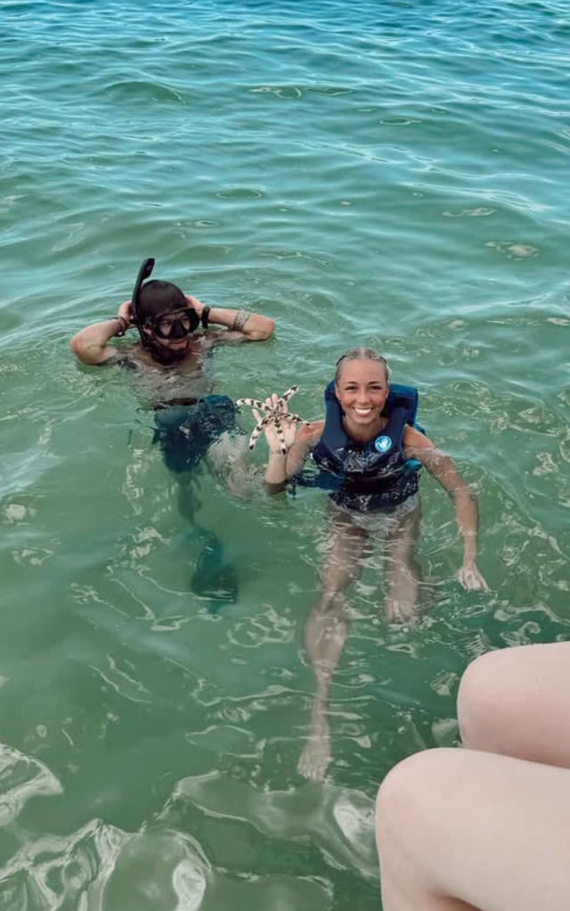 Two snorkelers in clear turquoise sea near a boat — smiling swimmer in a life vest holding a spotted starfish while a snorkeler adjusts a mask nearby.