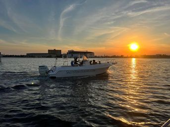 White motorboat with passengers cruising across a bay at golden sunset, sun reflecting on rippled water and a low waterfront skyline on the horizon.