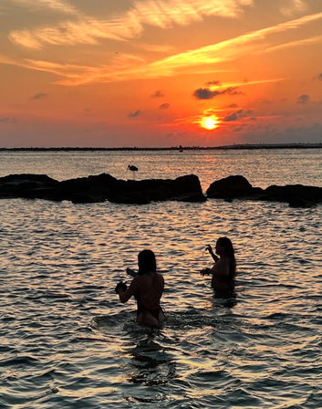 Two silhouetted people wading in shallow ocean water at a vibrant orange sunset, golden reflections on rippling sea and a shorebird perched on a rocky jetty