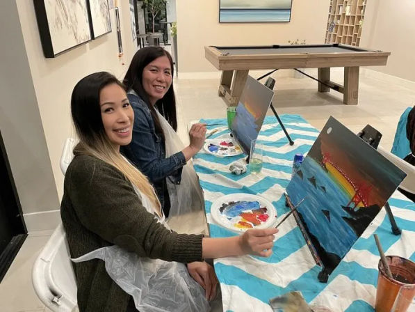 Two participants at an indoor paint-night table with a blue and white striped tablecloth, smiling as they paint sunset seascape canvases with palettes and brushes, a pool table visible in the background.
