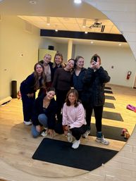 Eight women taking a mirror selfie after a yoga/fitness class in an indoor studio, smiling on black exercise mats on a light wood floor with ceiling lights and studio equipment in the background.