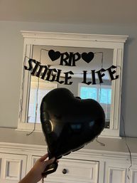 Black heart-shaped balloon held in front of a white dresser and mirror decorated with a black "RIP Single Life" banner — indoor bachelorette/bridal party decor.