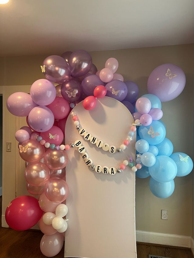 Indoor home party backdrop with a pastel balloon arch in pink, purple and blue, butterfly decals and a personalized letter-bead garland on a white arched panel — whimsical pastel party decor.
