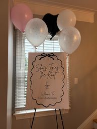 Playful indoor bachelorette welcome sign on an easel in a home entryway, surrounded by pink, white and black balloons; sign features a bow frame, script "She's Tying The Knot" and a small champagne glass icon.