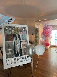 Bridal shower decor in a home: easel with a large photo-collage sign celebrating the bride-to-be, silver and white helium balloons in front, pastel pink balloon arch and hanging holographic butterfly garlands in a sunlit living room with hardwood floors.