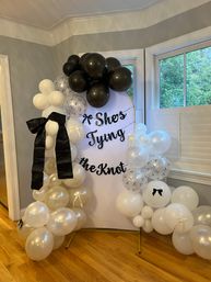 Indoor bridal shower backdrop in a home with a white sign reading She's Tying the Knot, surrounded by black, white and pearl balloons, confetti balloons and a large black bow on a hardwood floor near windows.