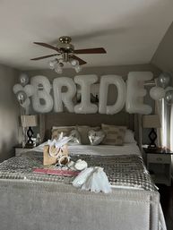 Bridal bedroom setup with oversized white letter balloons spelling "BRIDE" above a bed, silver heart and helium balloons, gift bag, veil and sash arranged on a tufted bedspread.