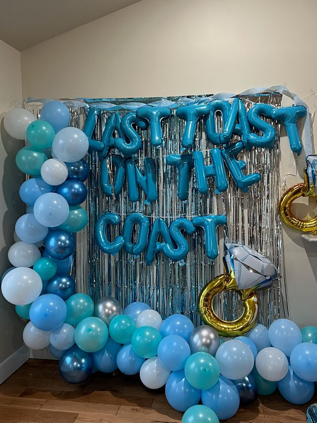 Silver fringe backdrop decorated with teal, aqua, sky-blue, white and metallic balloons, blue letter foil balloons spelling a celebratory message, and a gold ring-shaped foil balloon with a diamond — festive engagement or party backdrop.