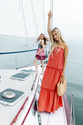 Two women on a sailboat deck at sea, one in a flowing red maxi dress holding a straw tote and the rigging, the other in a patterned mini dress near the mast against a calm, overcast ocean.