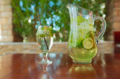 Refreshing mint-and-lime pitcher with ice and lime slices next to a drinking glass and straw on a wooden patio table, blurred green garden background — bright summer outdoor drink scene.