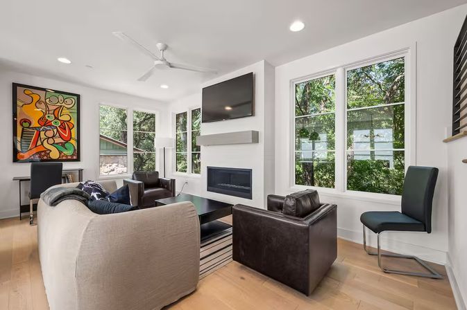 Bright modern living room with hardwood floors, large windows overlooking leafy trees, wall-mounted TV above a sleek fireplace, neutral sofa, leather armchairs, abstract wall art and ceiling fan