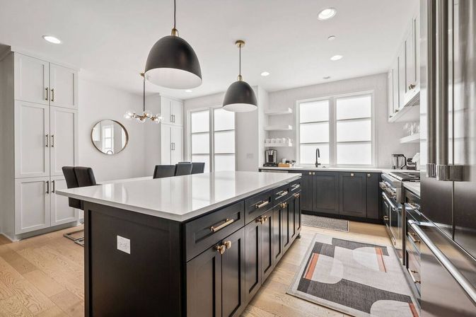 Bright modern kitchen with large white quartz island, dark shaker cabinets and brass hardware, black dome pendant lights, stainless appliances, and light oak floors by sunlit windows