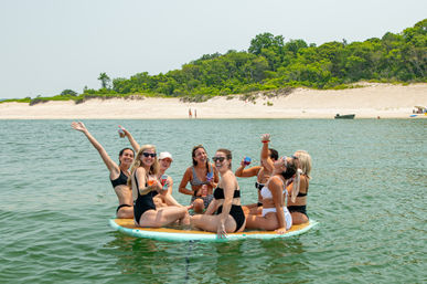 Eight women in swimsuits laughing and raising drinks while sitting on a large paddleboard in calm coastal water near a sandy beach with green dunes — summer beach paddleboard party.