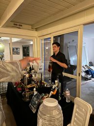 Home bartender serving drinks at a cocktail station on an enclosed patio — table stacked with liquor bottles, mixers and coolers, a guest’s hand reaching toward the bar by sliding glass doors