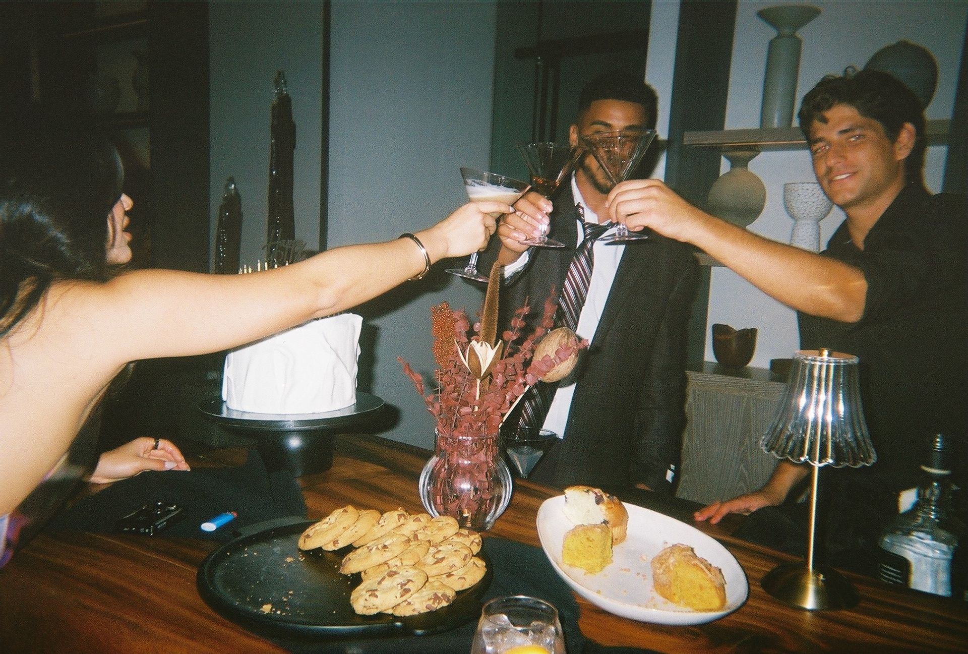 Group toasting with martini glasses at an indoor cocktail party over a wooden table with cookies on a black platter, sliced cake on a white plate, a white frosted cake on a stand, and a dried-flower centerpiece.