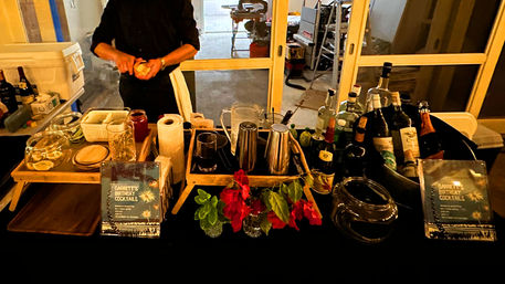 Mobile cocktail bar at a birthday party — bartender prepping drinks behind a black-clothed table with spirits, shakers, pitchers, garnish jars, fresh flowers and printed cocktail menus by sliding glass doors.