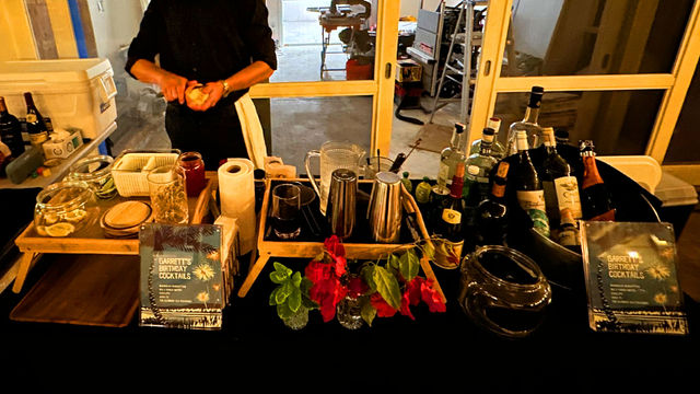 Mobile cocktail bar at a birthday party — bartender prepping drinks behind a black-clothed table with spirits, shakers, pitchers, garnish jars, fresh flowers and printed cocktail menus by sliding glass doors.