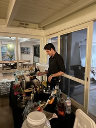 Bartender-style server mixing cocktails at an indoor home bar by sliding glass doors, table crowded with liquor bottles, mixers and glassware, coastal-themed dining area visible in background.