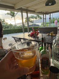 Hand holding an amber cocktail with a large ice cube on a covered tropical patio overlooking palm trees, ocean and a sunset; dining table with floral centerpiece and bottles in foreground.