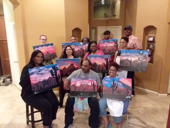Paint-night group of adults in a warm indoor room holding desert-sunset cactus paintings, smiling and posing with matching pink-and-blue mountain canvases.