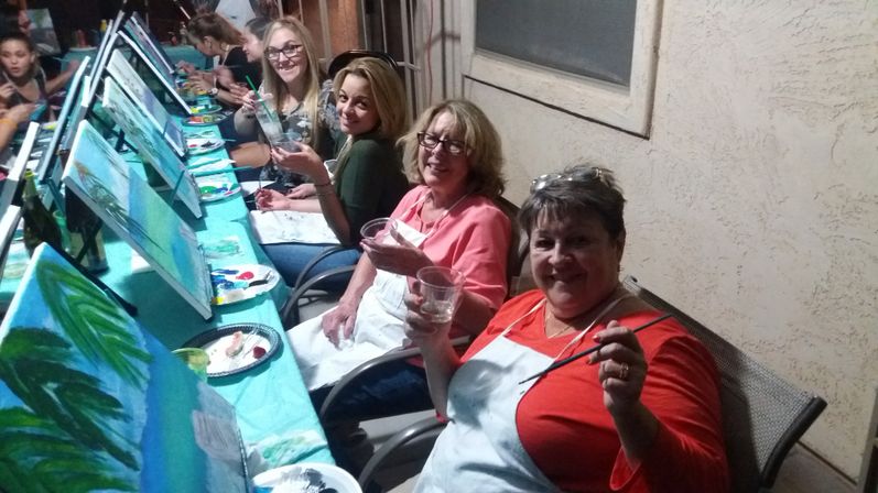 Group paint-and-sip party on a patio: smiling women in aprons seated along a long table, painting canvases on easels and holding brushes and drinks.