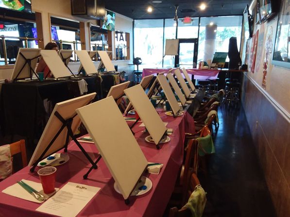 Blank canvases on tabletop easels and paint palettes lined up on pink-covered tables in a cozy art studio, ready for a group paint-and-sip painting class.