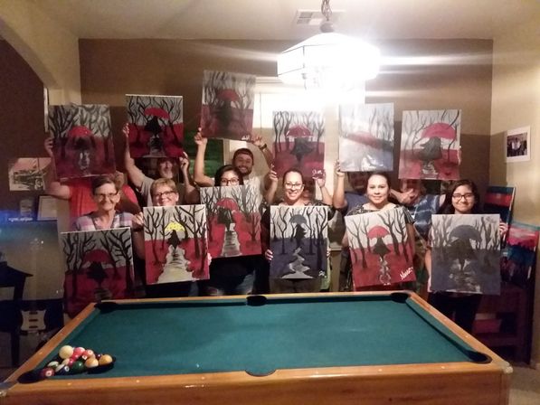 Cozy indoor paint night in a living room, a group poses behind a pool table holding matching canvases showing silhouetted figures with umbrellas in red-and-gray forest scenes.