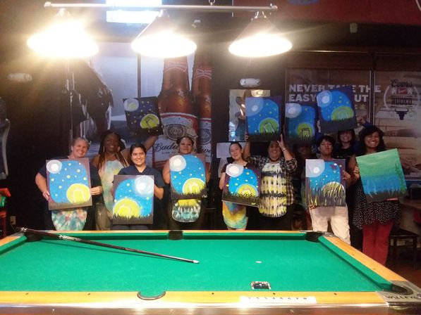 Smiling group at a paint-and-sip night in a bar, each holding a moonlit landscape canvas above a green pool table under three hanging lights.