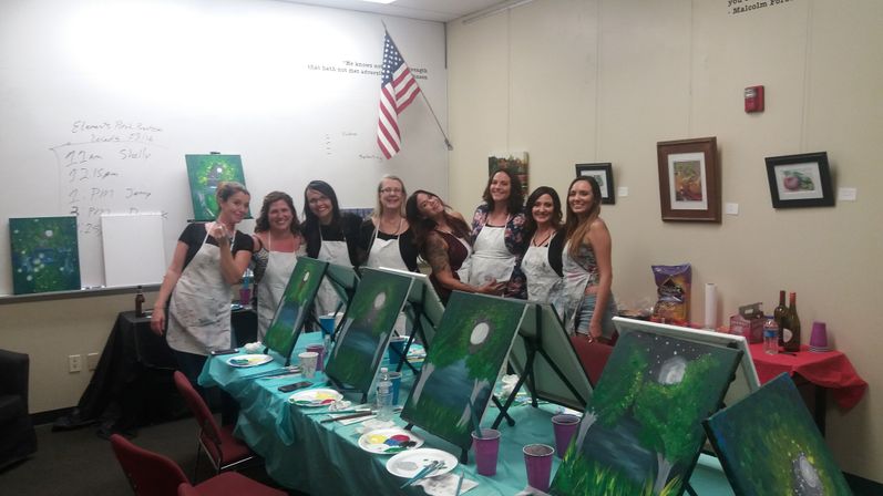 Group painting party in a classroom: cheerful women in aprons pose behind easels with green, moonlit lake paintings, paint palettes and cups on a covered table, American flag and framed art on the wall.