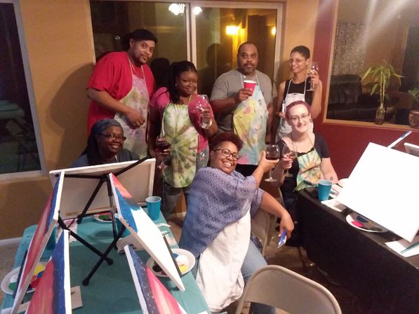 Seven adults at an indoor paint-and-sip party in a living room, wearing aprons and holding wine glasses around canvases, easels, and paint supplies.