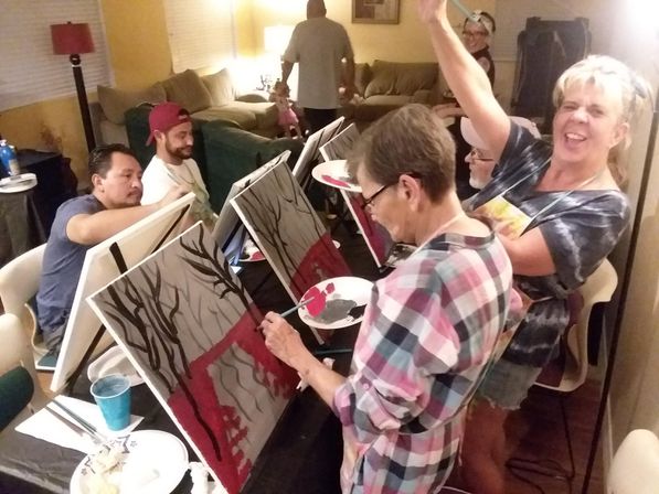 Cozy at-home paint night in a living room with friends at easels painting stylized tree silhouettes and red backgrounds, laughing and holding palettes.