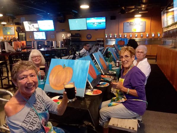 Group of women at an indoor paint-and-sip craft night in a bar, smiling and holding drinks while painting pumpkin canvases at a long table with beer taps and TV screens in the background.