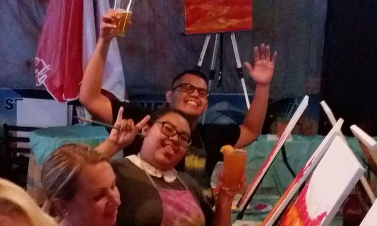 Three people at a paint-and-sip class indoors, smiling and raising drinks beside colorful canvases on easels