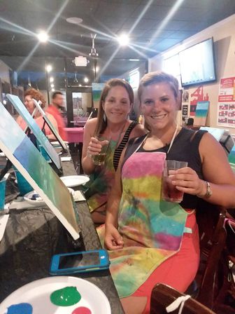 Two smiling women in colorful aprons holding drinks at a lively paint-and-sip class, canvases on easels and paint palette on the table in an art studio.